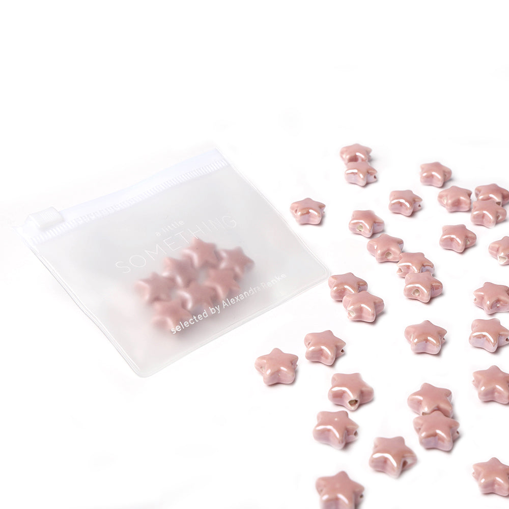 Rosafarbene Sternform-Perlen und glasierte Keramikperlen von Alexandra Renke sind in einem gefrosteten Reißverschlussbeutel auf weißem Hintergrund verstreut - ideal für die kreative Schmuckherstellung.
