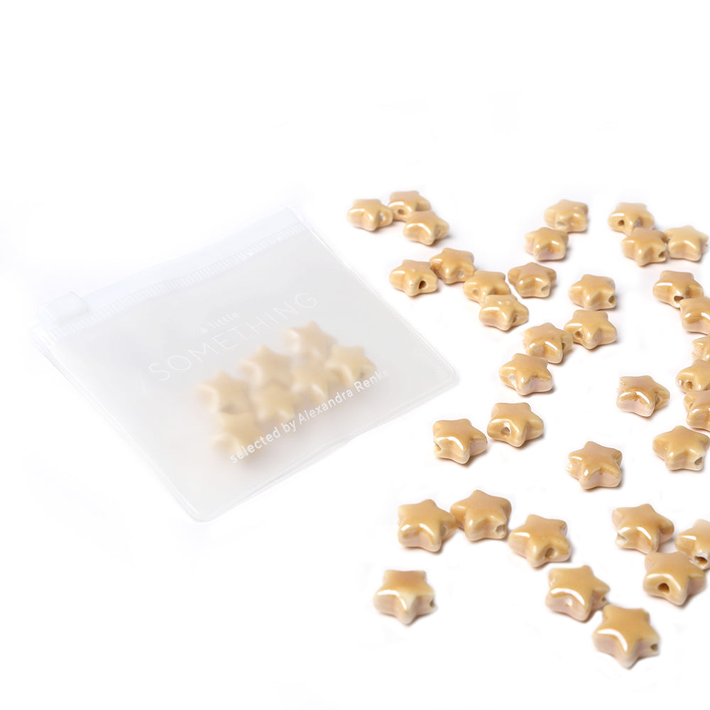 Kleine beige Sternform Perlen der Marke Alexandra Renke, perfekt für Schmuckdesign, liegen neben einem transparenten Zip-Beutel auf weißem Hintergrund verstreut.