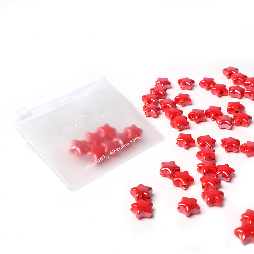 Ein durchsichtiger Beutel mit Alexandra Renke glasierten Keramikperlen in Sternform verstreut auf weißem Hintergrund - perfekt für kreative Schmuckgestaltung.