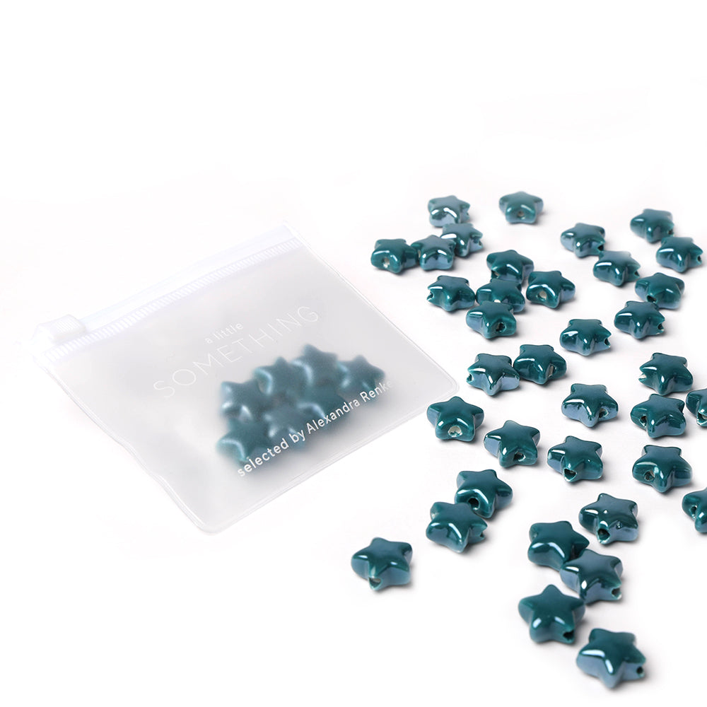 Die teal Sternform glasierten Keramikperlen von Alexandra Renke sind auf weißem Hintergrund neben einem halbtransparenten Beutel verstreut - ideal zur kreativen Schmuckgestaltung.