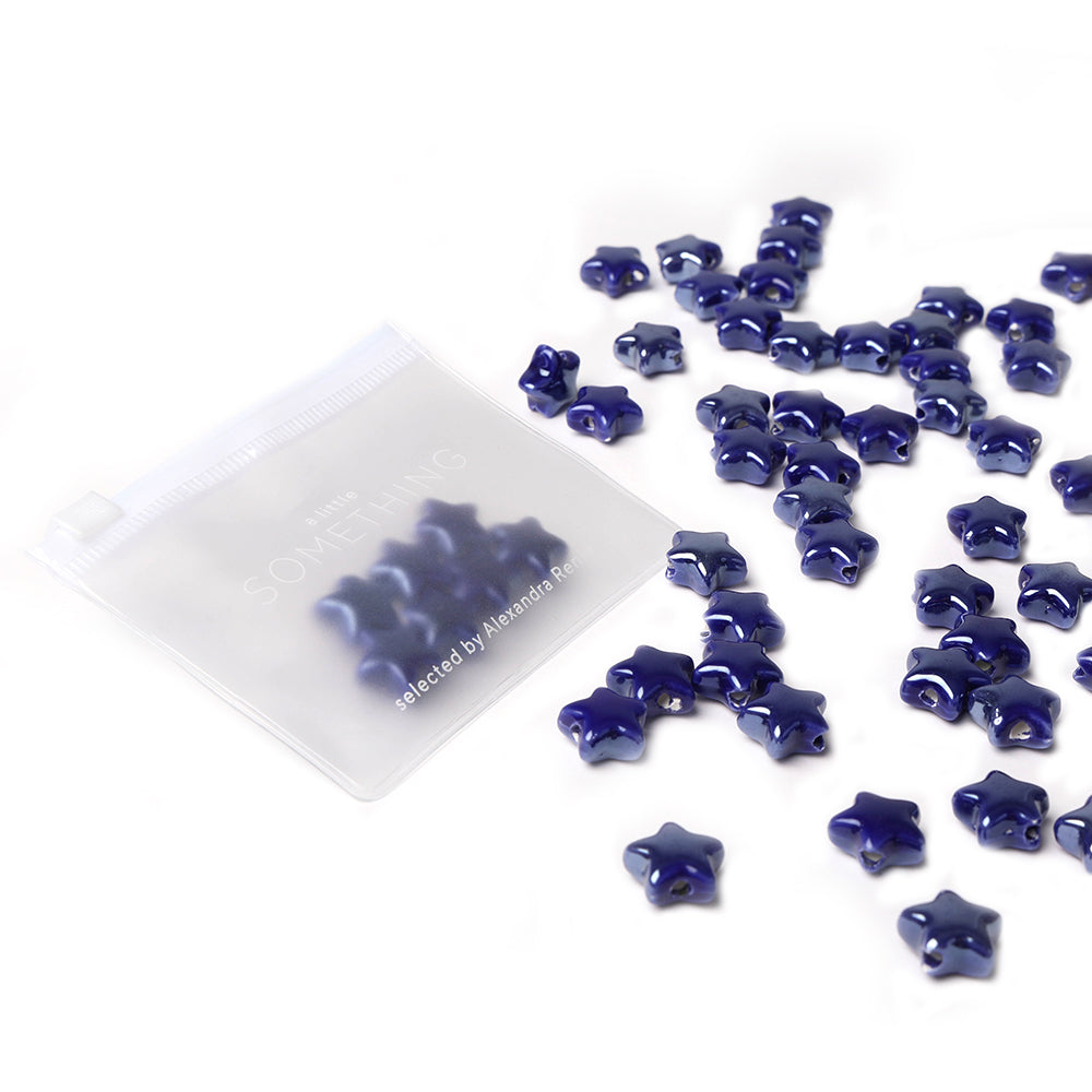 Kleine glänzende blaue Sternform-Perlen von Alexandra Renke, perfekt für die Schmuckherstellung, verstreut neben einem durchsichtigen Reißverschlussbeutel auf weißem Hintergrund.