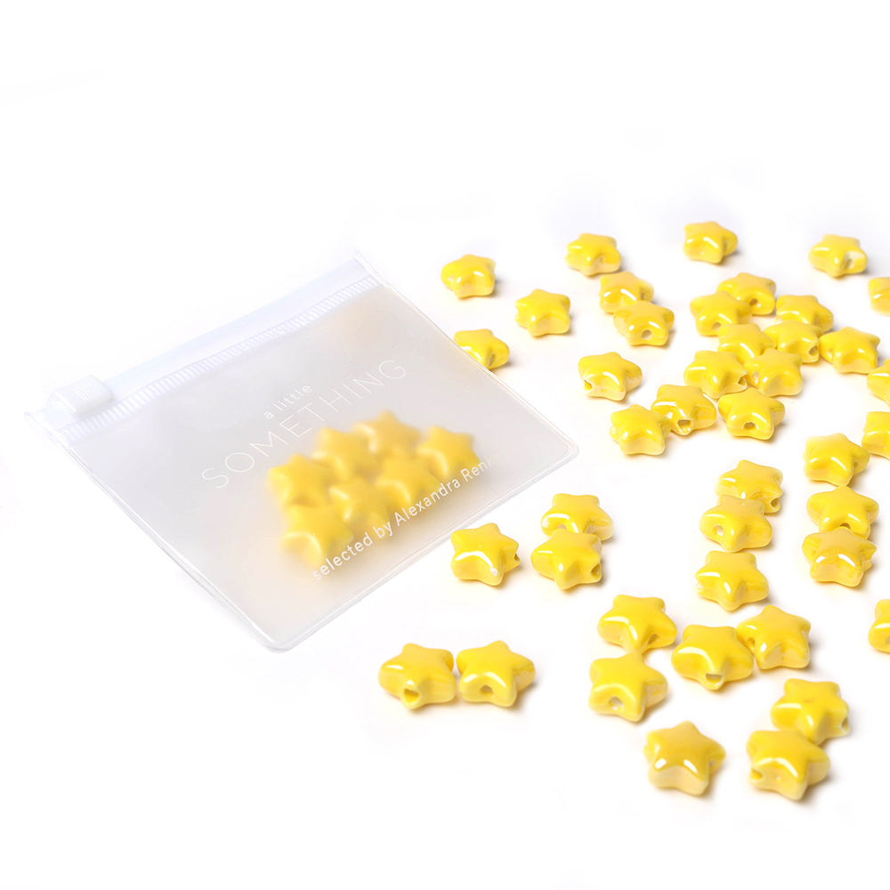 In einem durchsichtigen Beutel befinden sich gelbe Alexandra Renke Sternform Keramikperlen, daneben sind weitere glasierte sternförmige Perlen verstreut.