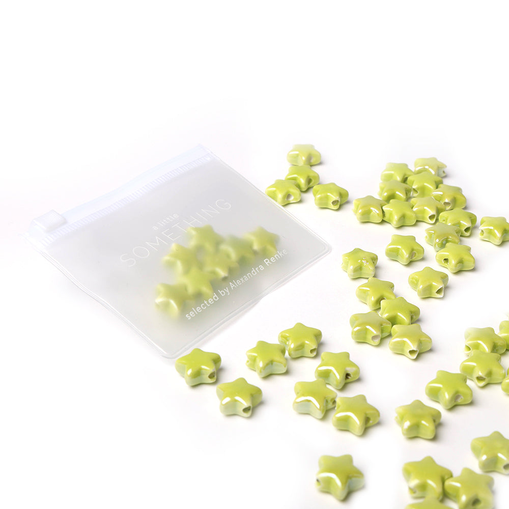 Kleine grüne, sternförmige, glasierte Keramikperlen von Alexandra Renke quellen aus einem durchsichtigen Beutel auf weißem Grund - ideal für die Schmuckherstellung.