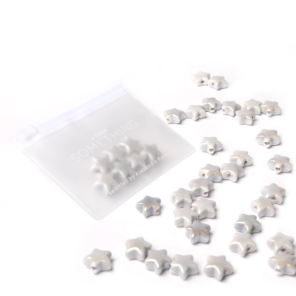 Kleine silberne Sternform-Perlen von Alexandra Renke sind neben einem durchsichtigen Reißverschlussbeutel auf einer weißen Fläche verstreut, ideal für Schmuckherstellung-Projekte.