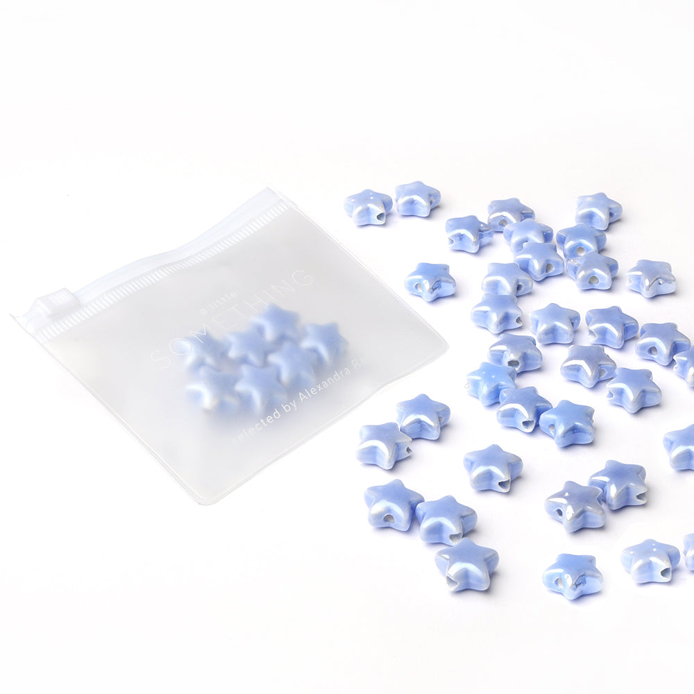 Die hellblau glasierten Keramik-Sternperlen von Alexandra Renke liegen verstreut neben einem transparenten Reißverschlussbeutel auf weißem Grund - ideal für kreative Schmuckprojekte.