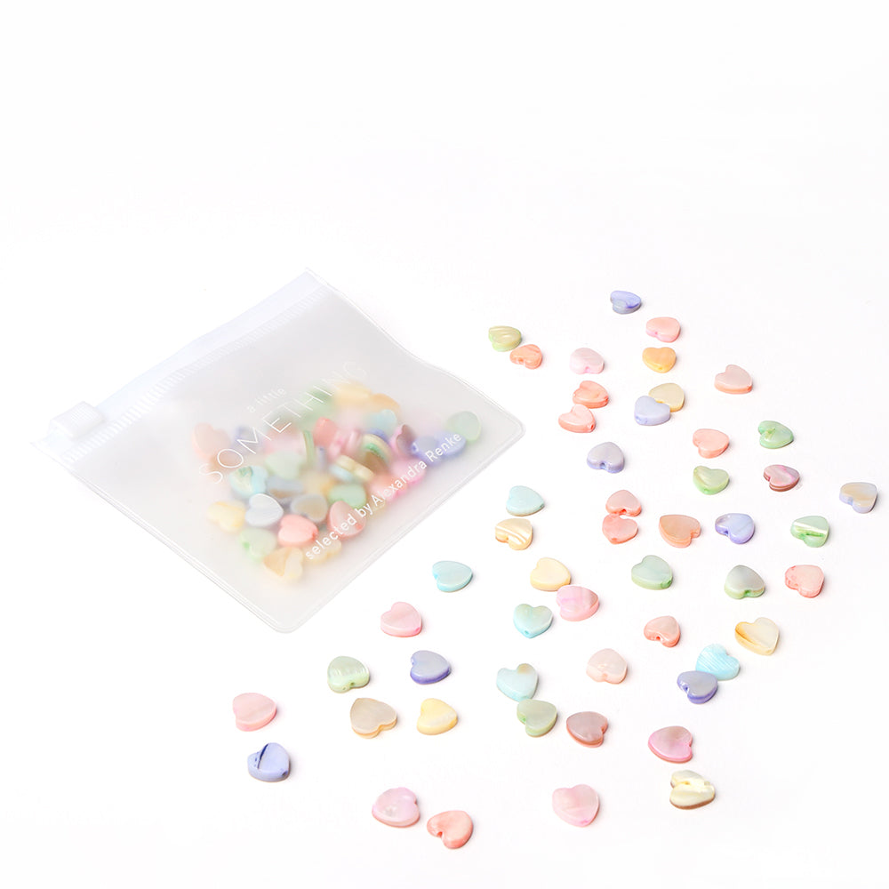 Kleine pastellfarbene Herzperlen von Alexandra Renke, darunter Muschelperlen Herzform, befinden sich neben einem durchsichtigen Reißverschlussbeutel auf weißem Grund - ideal zum Schmuck basteln oder als Ergänzung zu Ihrem Lieblings-Perlen-Mix.