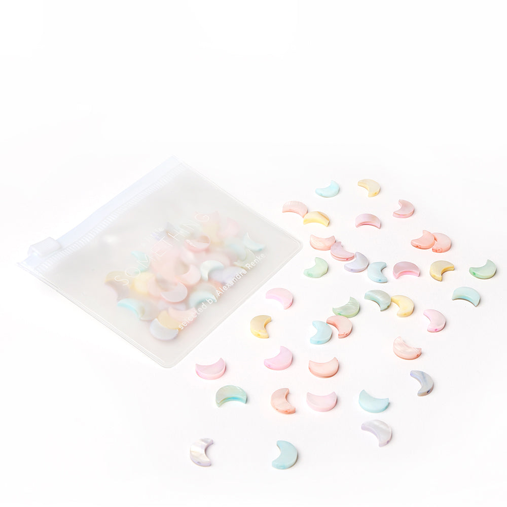 Ein durchsichtiges Täschchen mit verstreuten pastellfarbenen Muschelperlen in Mondform von Alexandra Renke - perfekt für Schmuck basteln oder Perlen Anhänger - auf weißem Hintergrund.