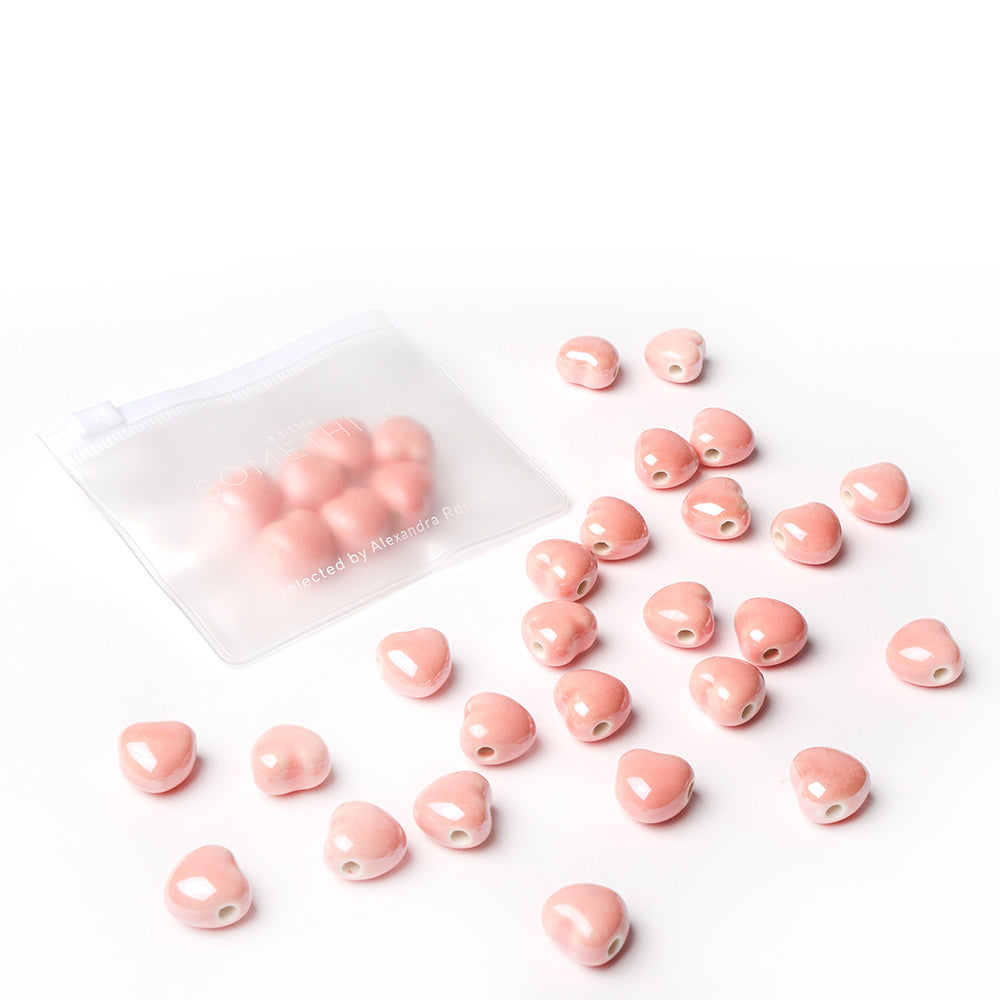Kleine rosa glasierte Keramikperlen von Alexandra Renke verstreut neben einem gefrosteten Reißverschlussbeutel auf weißem Hintergrund - ideal für Schmuck basteln und kreative Schmuckdesigns.