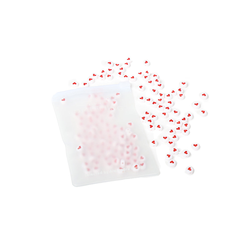 Ein ☆ Beutel, der mit kleinen runden Perlen gefüllt ist, die jeweils mit einem roten Herzmotiv verziert sind und auf einem weißen Hintergrund verstreut liegen - ideal für die Herstellung von Schmuck.