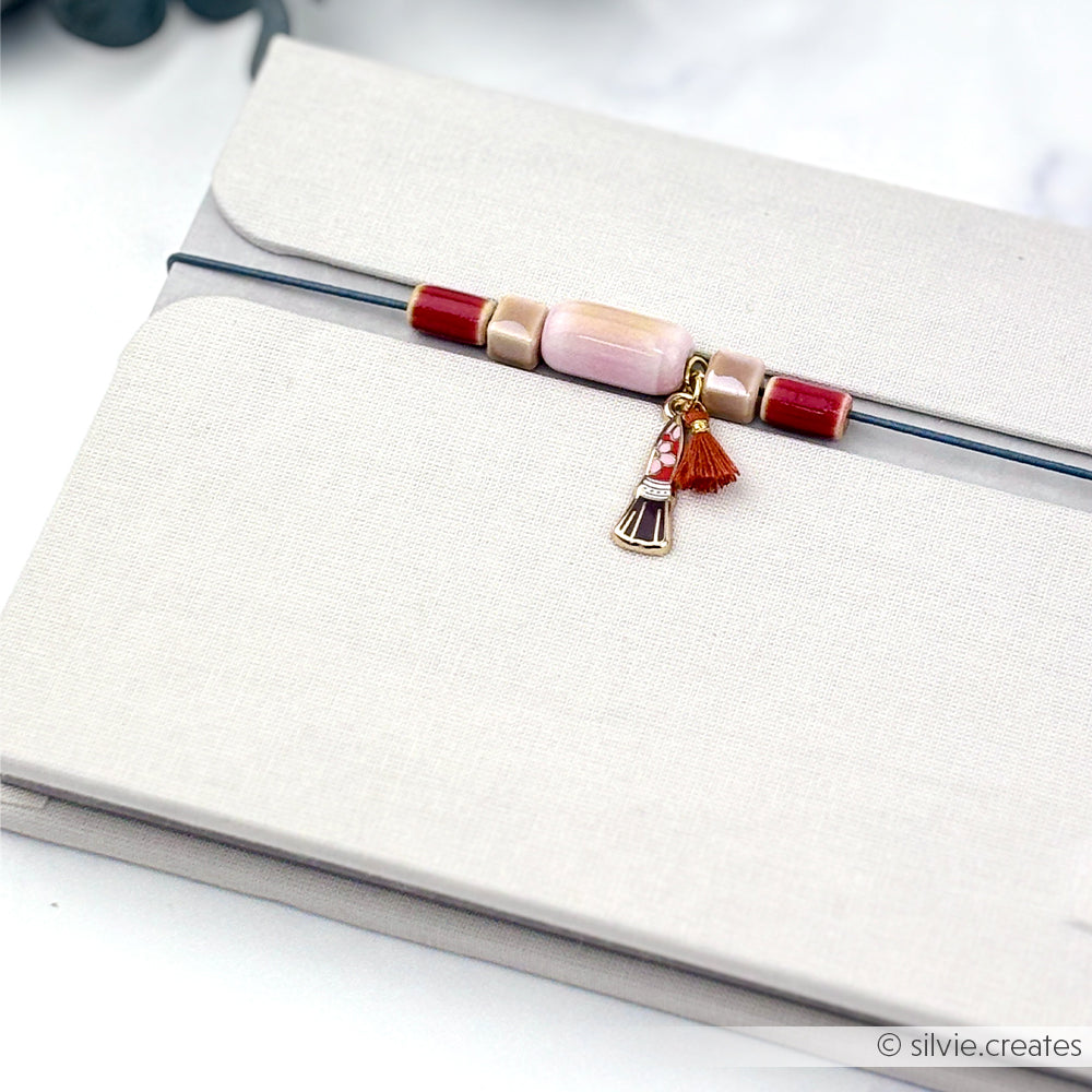Ein beiges Notizbuch mit einem Perlenanhänger und einer Quaste an einem Gummiband, verziert mit glasierten Keramikperlen – die perfekte Inspiration für alle, die gerne mit Perlen basteln und selbst Schmuck herstellen.