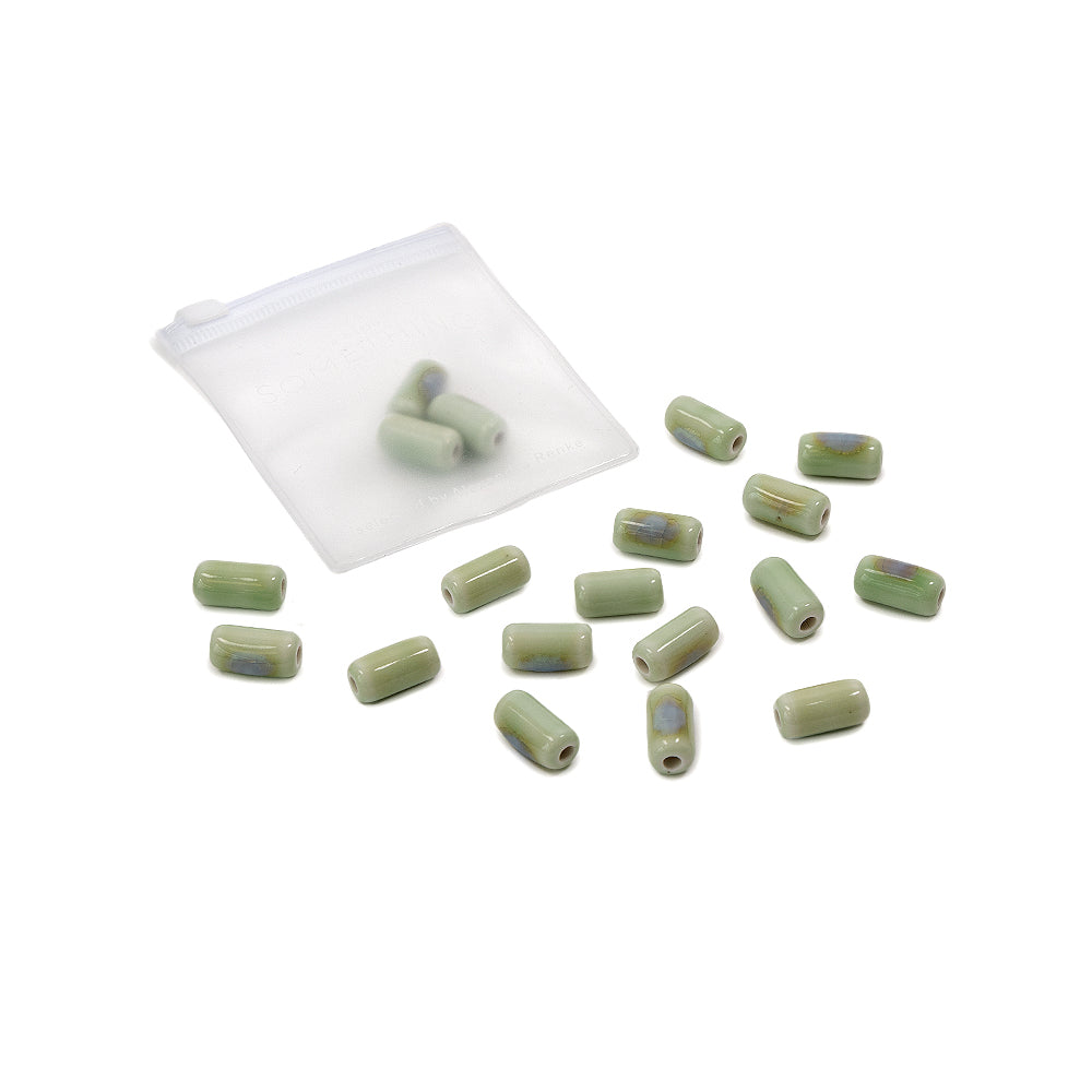 Kleine grüne, zylindrische Perlen, ideal zum Perlenbasteln und zur Herstellung von selbstgemachtem Schmuck, liegen verstreut neben einem durchsichtigen Reißverschlussbeutel auf weißem Hintergrund.