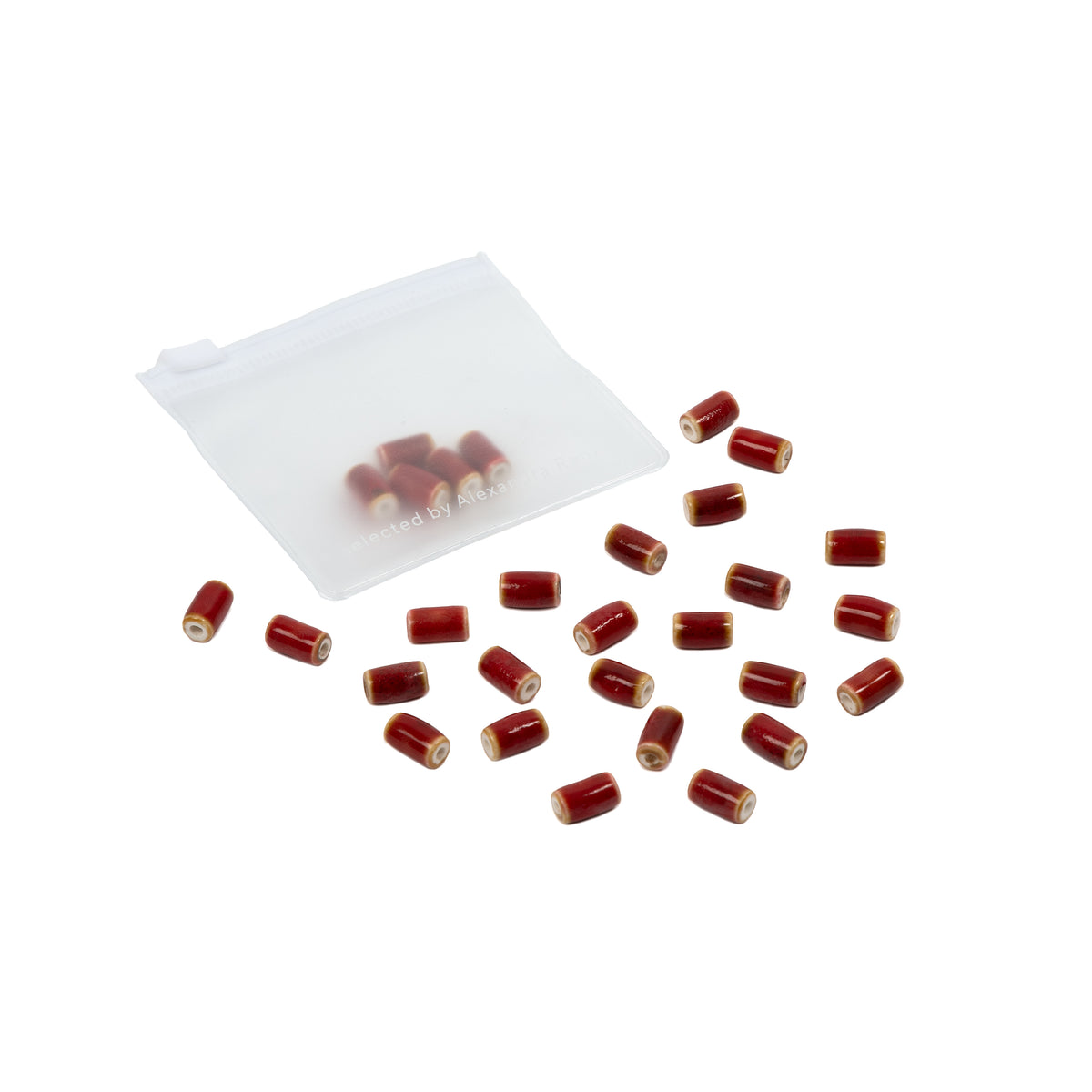 Kleine rote, zylindrische Perlen, die an handgefertigte Perlen erinnern, sind auf weißem Hintergrund neben einem durchsichtigen Reißverschlussbeutel verstreut – perfekt für alle, die gerne selbst Schmuck basteln.