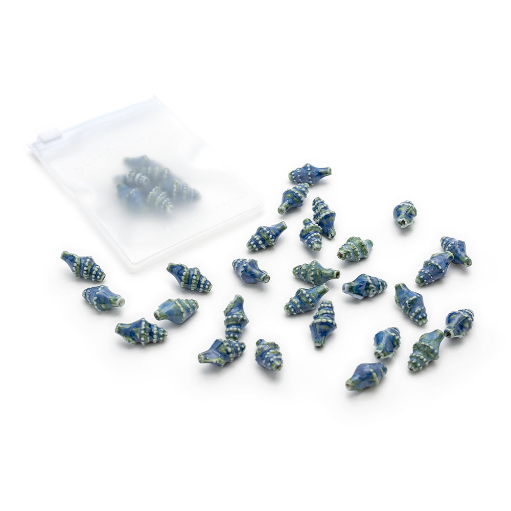 Kleine, blau und grün glasierte Keramikperlen in Muschelform von Alexandra Renke liegen verstreut neben einem durchsichtigen Beutel, der mit weiteren handgefertigten Perlen gefüllt ist.