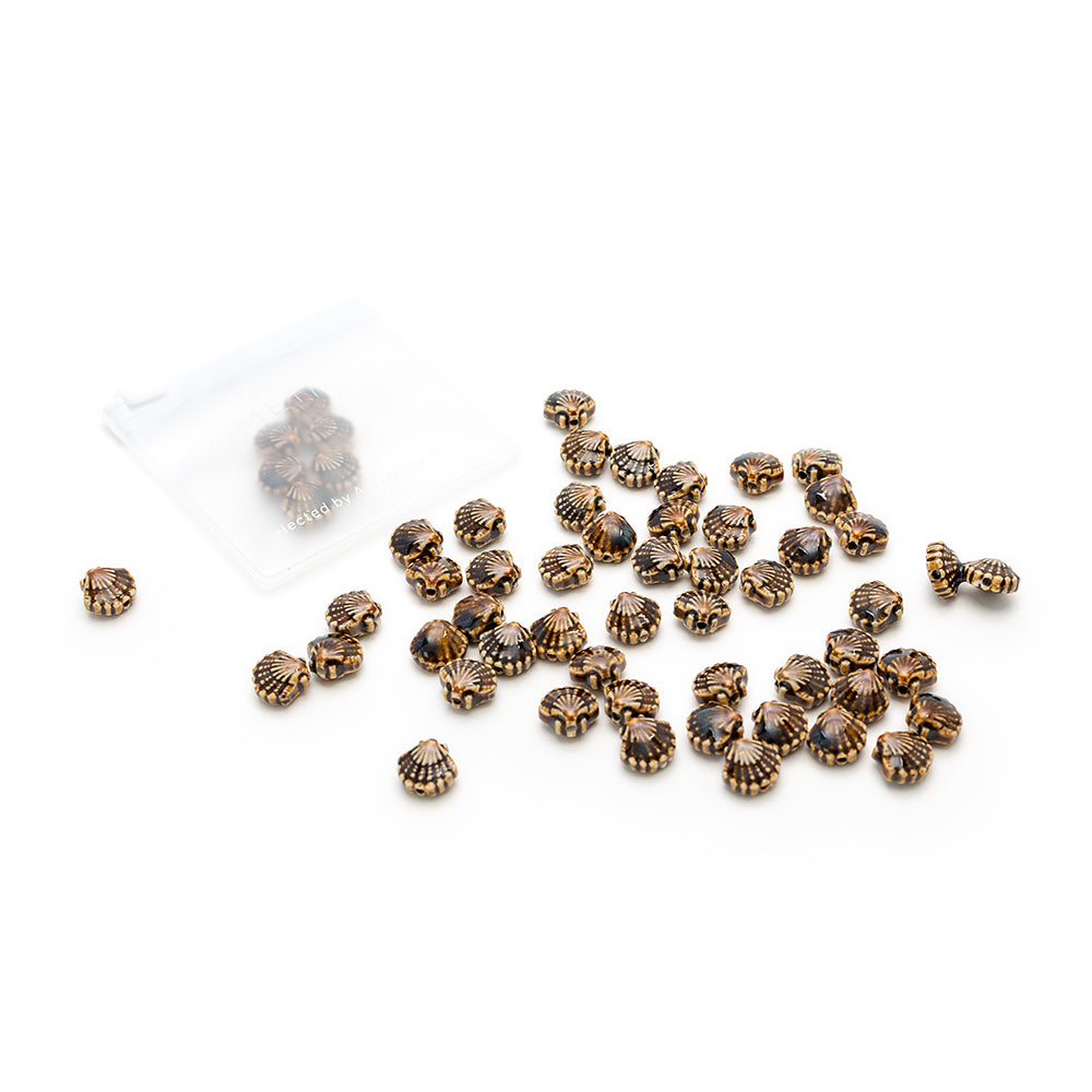 Kleine, runde, goldene und schwarze Perlen der Marke Alexandra Renke liegen auf weißem Hintergrund verstreut neben einem durchsichtigen Plastikbeutel.