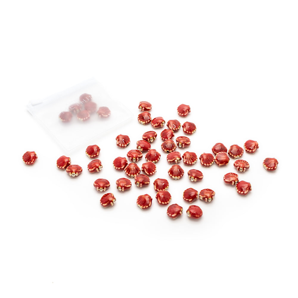 Auf einem weißen Hintergrund liegen verstreut rote Glasperlen von Alexandra Renke mit goldenen Akzenten sowie handgefertigte Perlen in Muscheloptik neben einem kleinen durchsichtigen Plastikbeutel.