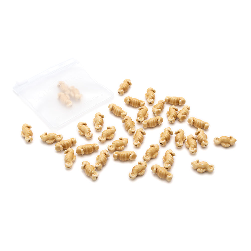 Kleine, beigefarbene Nudeln in Rotini-Form von Alexandra Renke, verstreut neben einem durchsichtigen Beutel auf weißem Hintergrund, erinnern an handgefertigte Perlen im Seepferdchen-Look oder glasierte Keramikperlen.