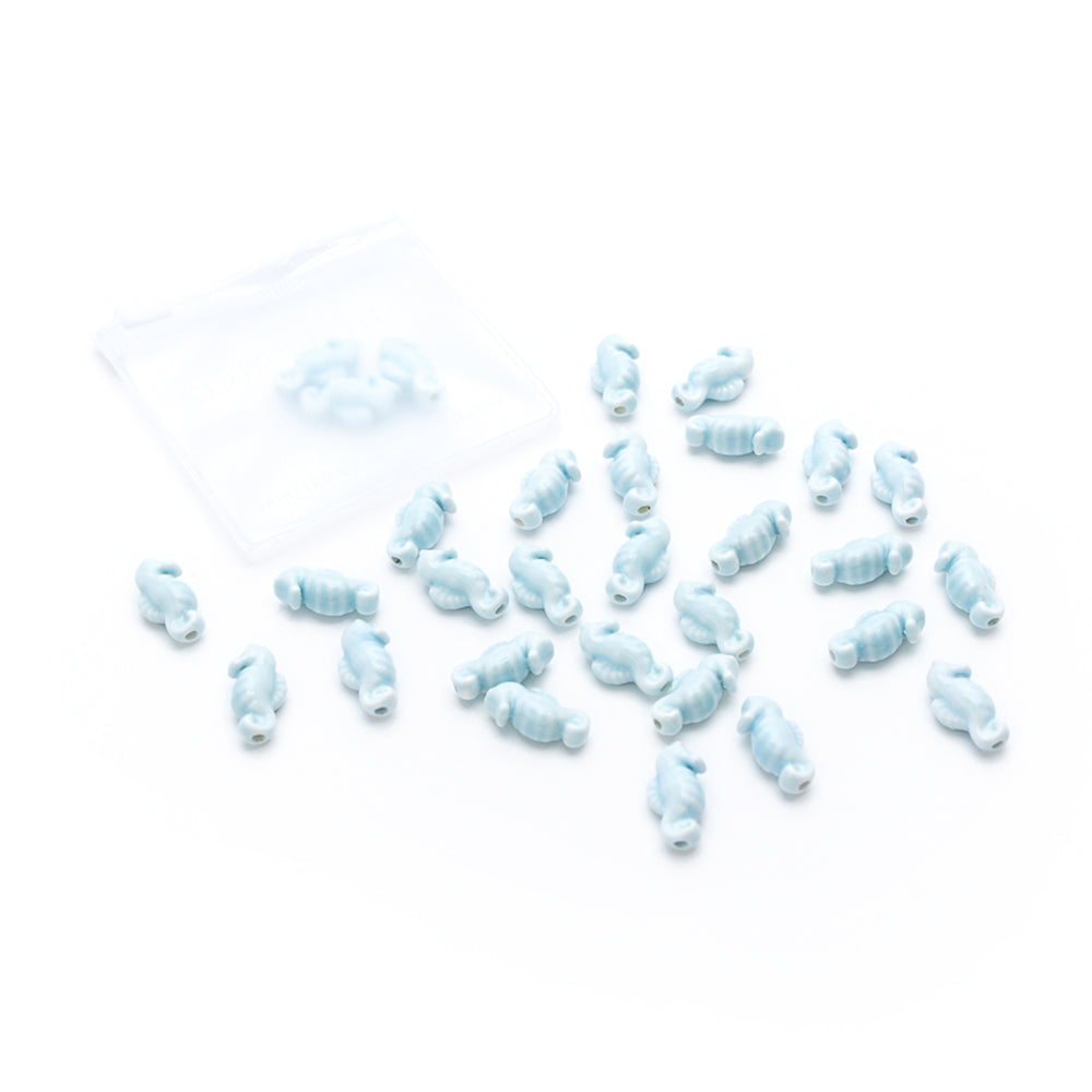 Mehrere kleine, hellblau glasierte Keramikperlen von Alexandra Renke liegen verstreut neben einer kleinen durchsichtigen Plastiktüte auf einem weißen Hintergrund.