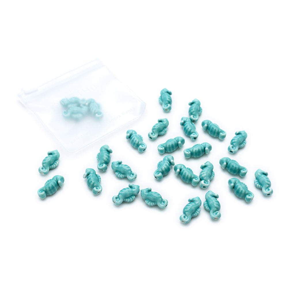 Kleine türkisfarbene Anhänger in Skorpionform von Alexandra Renke, inspiriert von der Seepferdchen-Optik, liegen verstreut neben einer durchsichtigen Plastiktüte auf weißem Hintergrund.
