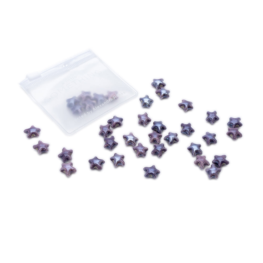 Auf weißem Hintergrund sind violette, glitzernde, sternförmige Objekte von Alexandra Renke in der Nähe eines kleinen durchsichtigen Beutels mit Reißverschluss verstreut, die an handgefertigte Keramikperlen erinnern.