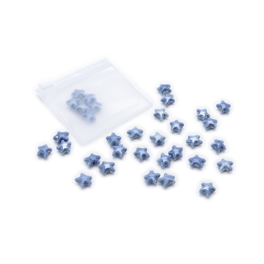 Kleine blaue handgefertigte Perlen im Seestern-Design der Marke Alexandra Renke sind auf weißem Hintergrund neben einem durchsichtigen Plastikbeutel verstreut.