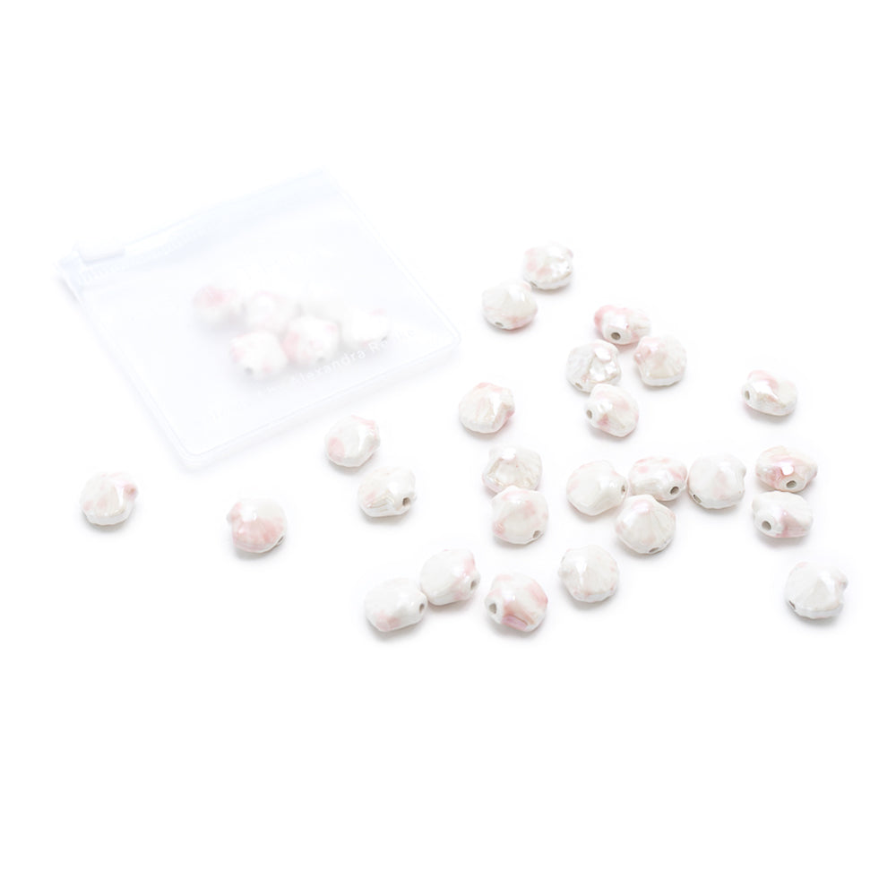 Kleine, runde, weiß-rosa marmorierte glasierte Keramikperlen von Alexandra Renke liegen verstreut auf einer weißen Fläche in der Nähe eines teilweise geöffneten Plastikbeutels mit Reißverschluss.