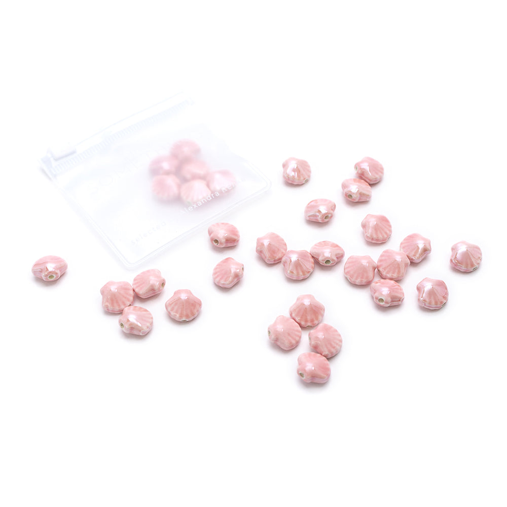 Handgefertigte, rosa Perlen in Muscheloptik von Alexandra Renke liegen verstreut auf einem weißen Hintergrund neben einem kleinen, durchsichtigen Beutel mit Reißverschluss.