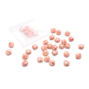 Kleine rosa Perlen in Muschelform von Alexandra Renke in Muschel-Optik sind auf einer weißen Fläche verstreut, wobei ein Netzbeutel einige der handgefertigten, glasierten Keramikperlen enthält.