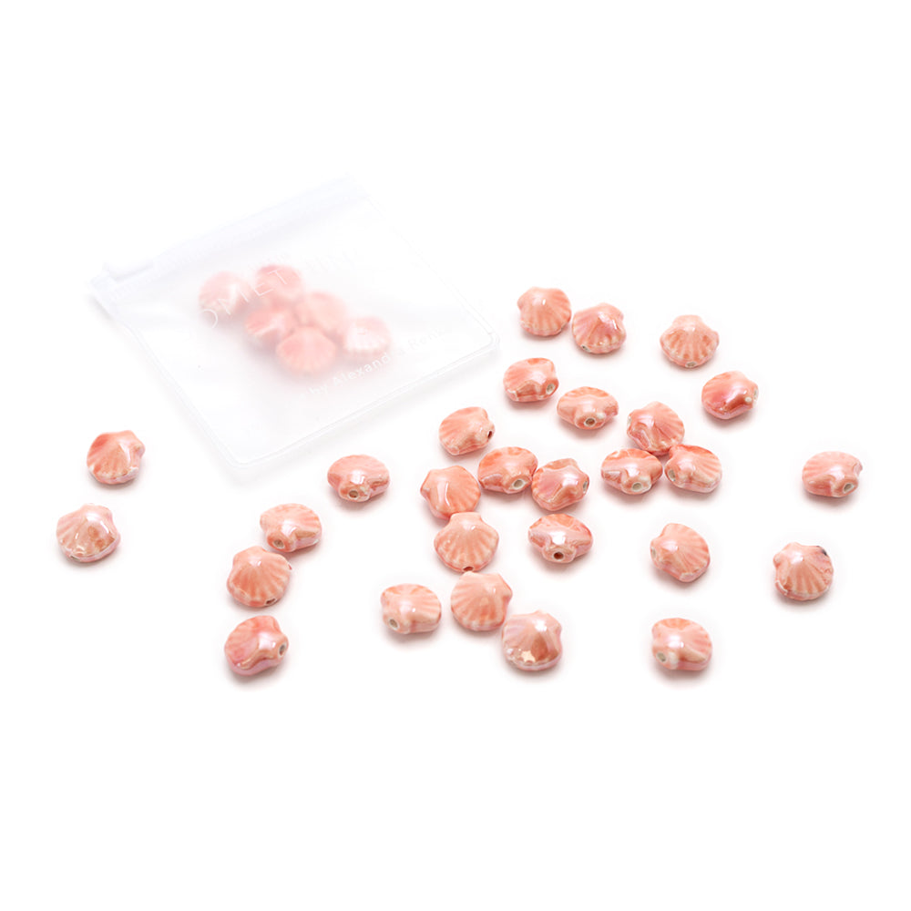 Kleine rosa Perlen in Muschelform von Alexandra Renke in Muschel-Optik sind auf einer weißen Fläche verstreut, wobei ein Netzbeutel einige der handgefertigten, glasierten Keramikperlen enthält.