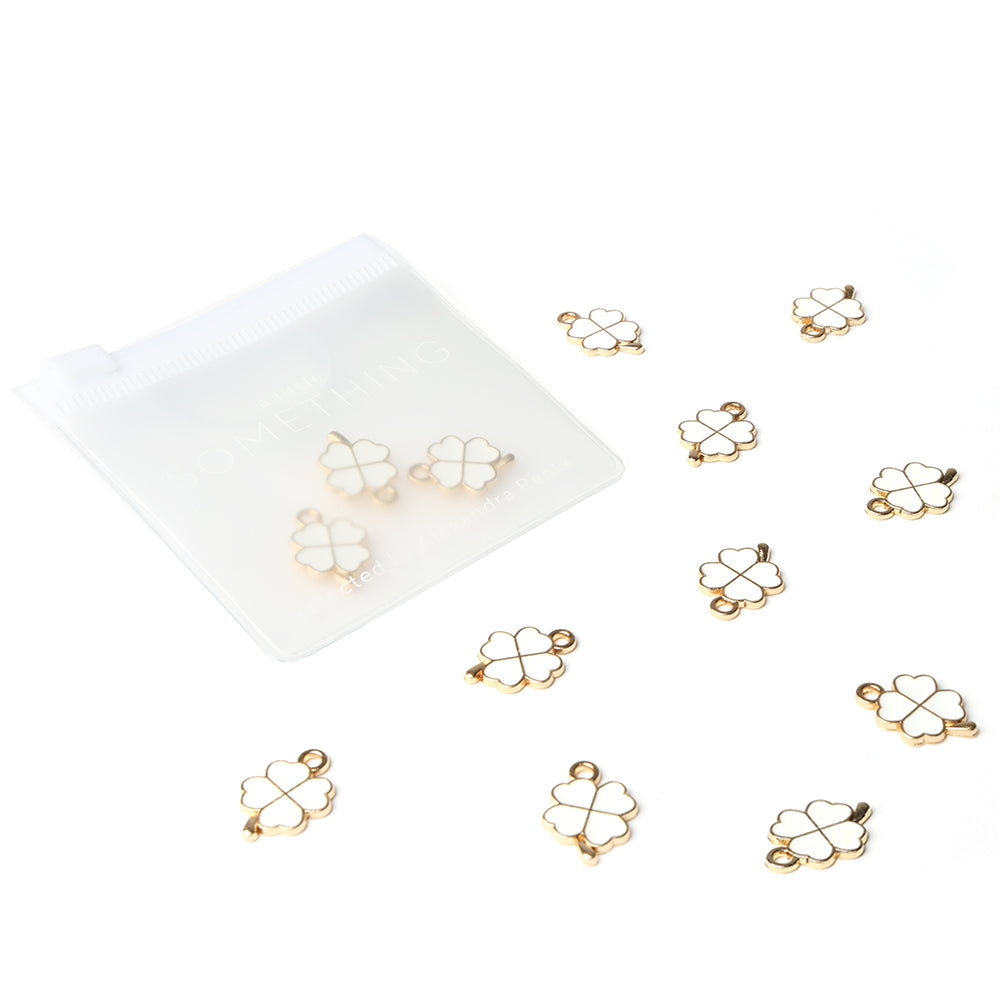 Alexandra Renke Gold und Weiß Glücksklee Charm Schmuckanhänger verstreut an einem durchsichtigen Beutel auf Weiß, ideal als Buchbinde Zubehör oder zur Schmuckherstellung.