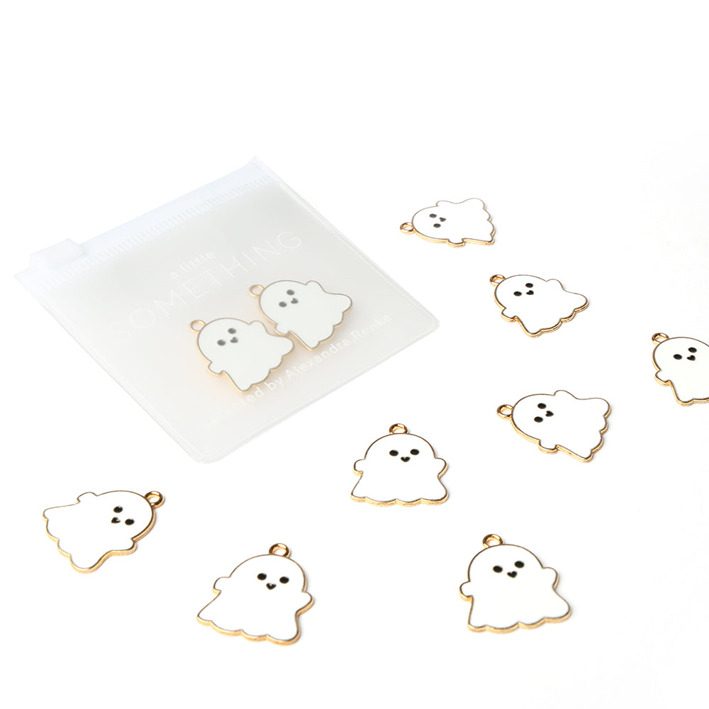 Acht kleine Schmuckanhänger in Geisterform von Alexandra Renke, goldfarben umrandet und in der Nähe eines gefrosteten Beutels angeordnet - ideal, um jedem Projekt einen gruseligen Buchbinden-Akzent zu verleihen.