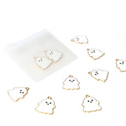 Acht kleine Schmuckanhänger in Geisterform von Alexandra Renke, goldfarben umrandet und in der Nähe eines gefrosteten Beutels angeordnet - ideal, um jedem Projekt einen gruseligen Buchbinden-Akzent zu verleihen.