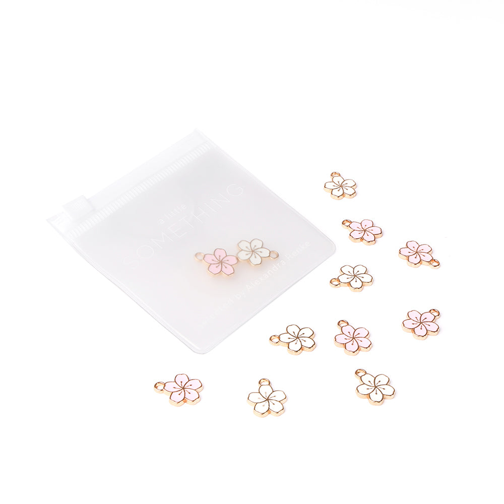 Die kleinen rosafarbenen und weißen Kirschblüten-Charm-Anhänger mit Goldrand von Alexandra Renke sitzen neben einem durchscheinenden weißen Reißverschlussbeutel - ideales Buchbindezubehör für Papierprojekte-Anhänger.