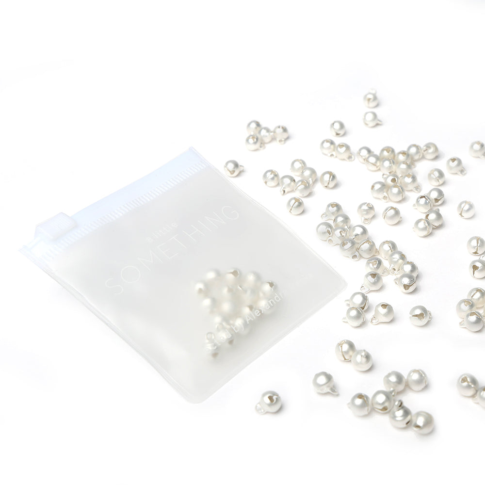 Die kleinen silbernen Glöckchen von Alexandra Renke sind neben einem durchsichtigen Kunststoffbeutel auf weißem Hintergrund zu sehen - ideal, um Ihre Scrapbooking-Projekte zu veredeln.