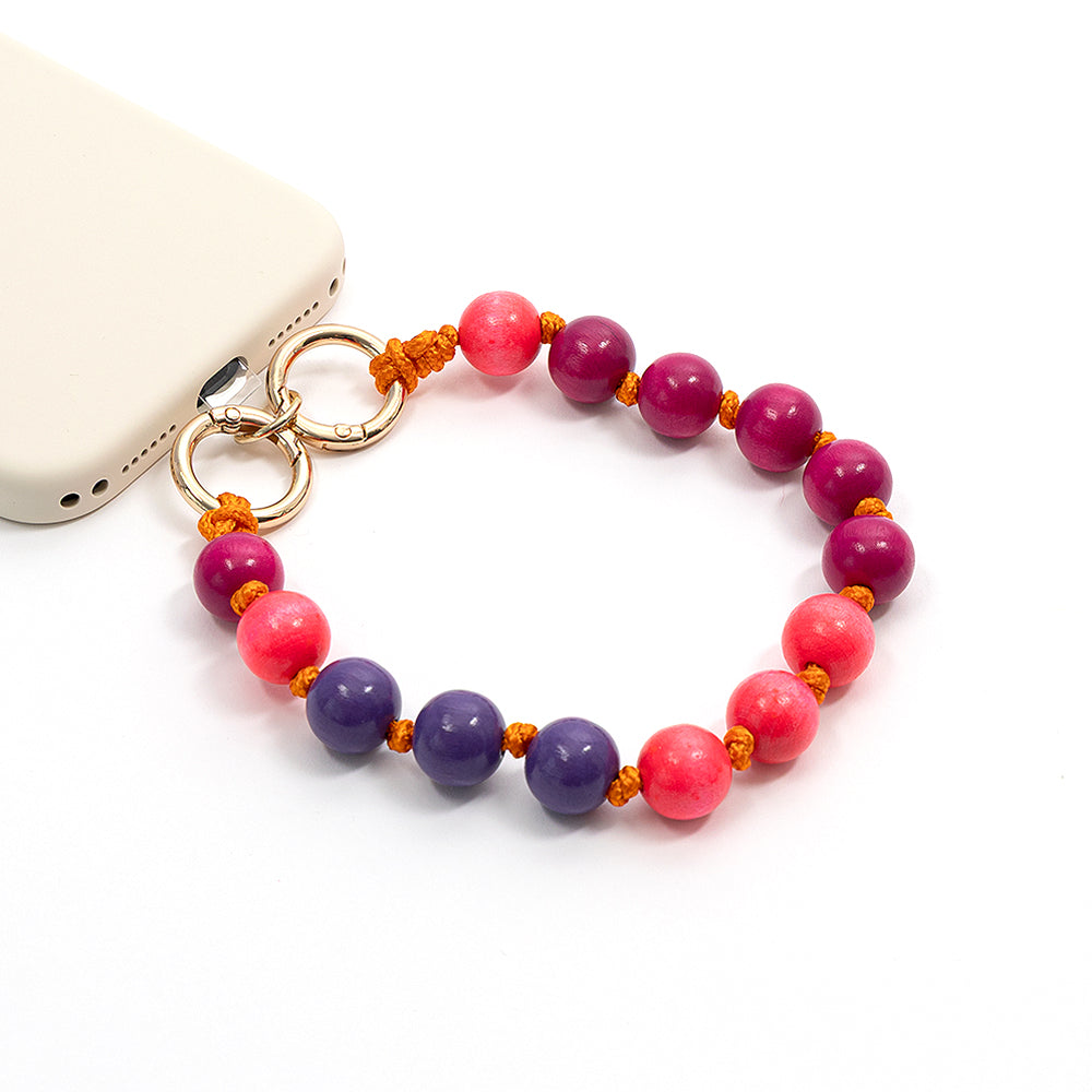 Ein Handy-Anhänger von Alexandra Renke aus rosa, lila und roten Perlen – darunter auch Holzperlen aus nachhaltigem Holz –, der mit goldenen Ringen an einem weißen Smartphone befestigt ist und sich somit perfekt als umweltfreundliches Bastelprojekt eignet, um dem Handy eine stilvolle Note zu verleihen.
