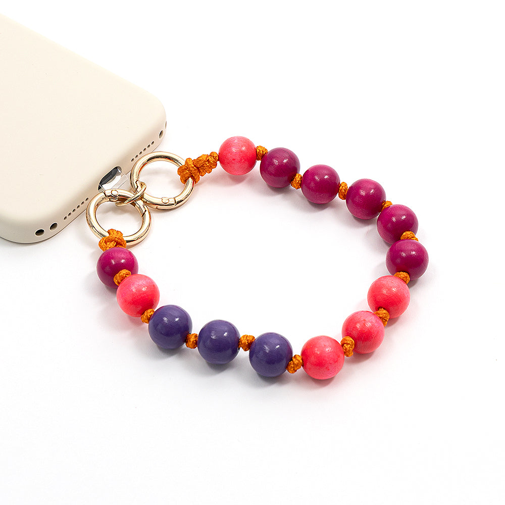 Ein Handy-Anhänger von Alexandra Renke mit violetten und rosa Perlen sowie Akzenten aus nachhaltigem Holz ist an einem beigen Handy vor weißem Hintergrund befestigt.