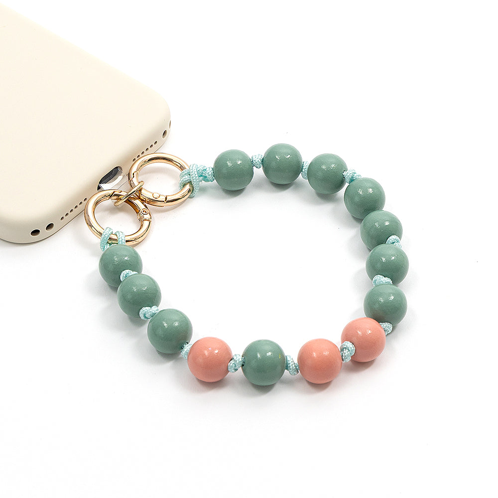 Beigefarbene Handyhülle von Alexandra Renke mit einer Handy-Schlaufe aus grünen und rosa Perlen, verziert mit einzigartigen Holzperlen und goldenen Ringen – ein perfektes Accessoire für alle, die gerne Schmuck basteln und den Look von nachhaltigem Holz lieben.