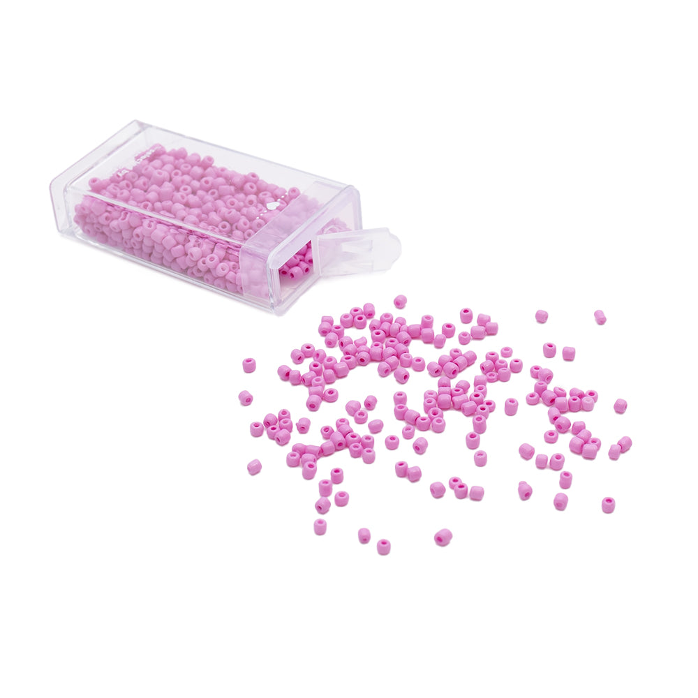 Eine durchsichtige Schachtel mit kleinen rosa Glasperlen von Alexandra Renke, von denen einige auf einer weißen Fläche verstreut sind – ideal, um Ihren Kreationen einen Vintage-Touch zu verleihen.