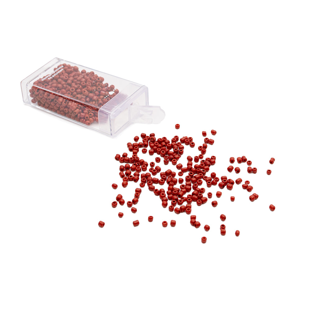 Kleine rote Rocailles-Perlen von Alexandra Renke liegen verstreut neben einem durchsichtigen Plastikbehälter auf weißem Hintergrund und verleihen dem Ganzen einen Hauch von Vintage-Charme.