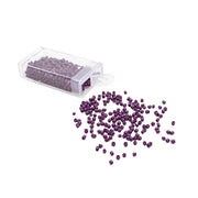 Eine durchsichtige Plastikschachtel mit kleinen violetten Rocailles-Perlen von Alexandra Renke, von denen einige auf einer weißen Fläche verstreut sind und so einen dezenten Vintage-Look erzeugen.