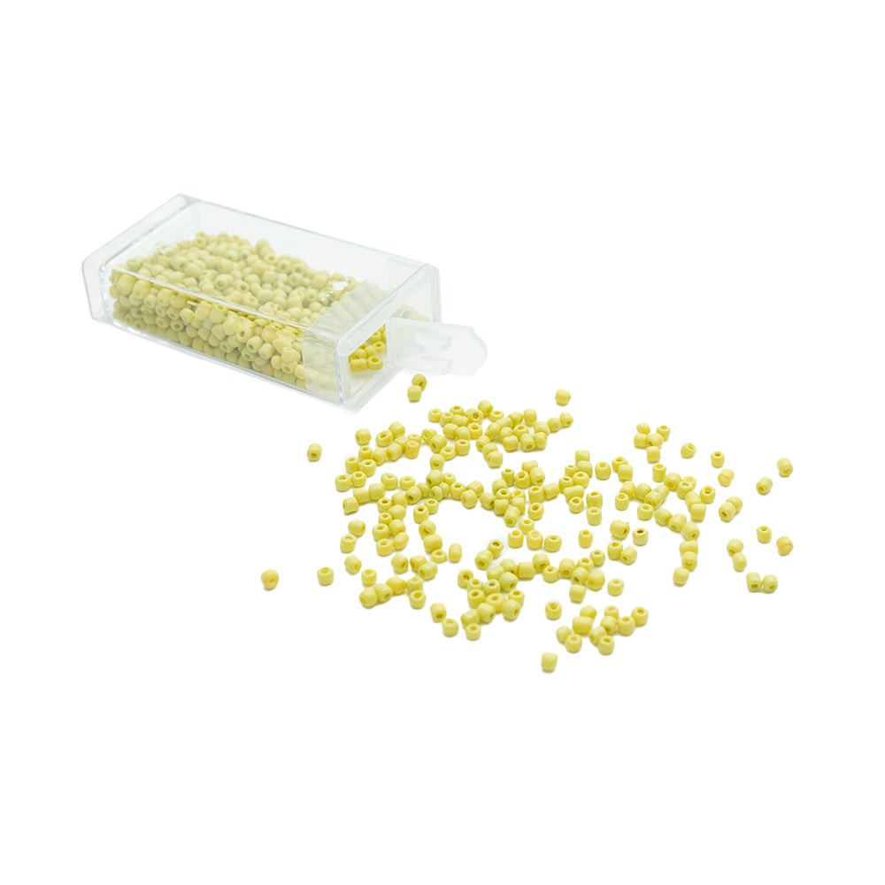Aus einem durchsichtigen Plastikbehälter mit kleinen gelben Rocailles-Perlen von Alexandra Renke ergießen sich die Perlen auf eine weiße Fläche und bringen so ihre klassische Glasperlen-Vintage-Optik besonders gut zur Geltung.
