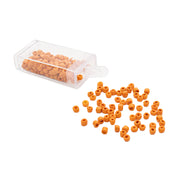 Kleine orangefarbene, zylindrische Rocailles-Perlen von Alexandra Renke, von denen einige aus einem durchsichtigen Plastikbehälter auf eine weiße Oberfläche gefallen sind, verleihen jedem Projekt den Charme einer Vintage-Optik.