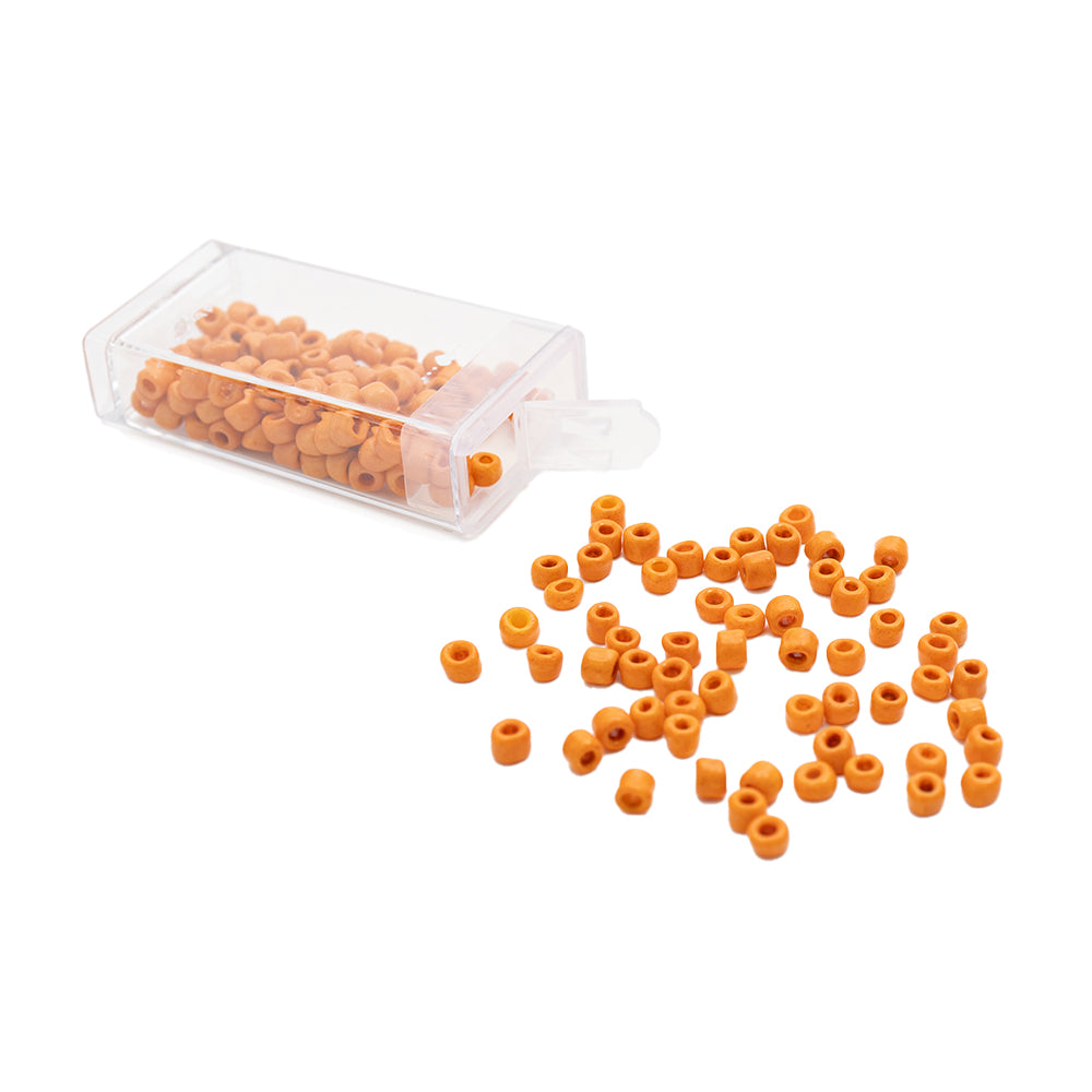 Kleine orangefarbene, zylindrische Rocailles-Perlen von Alexandra Renke, von denen einige aus einem durchsichtigen Plastikbehälter auf eine weiße Oberfläche gefallen sind, verleihen jedem Projekt den Charme einer Vintage-Optik.