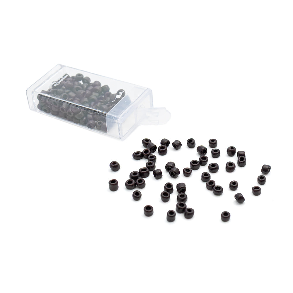 Kleine schwarze Rocailles-Perlen von Alexandra Renke im Vintage-Stil liegen verstreut neben einem durchsichtigen Plastikbehälter auf weißem Hintergrund.