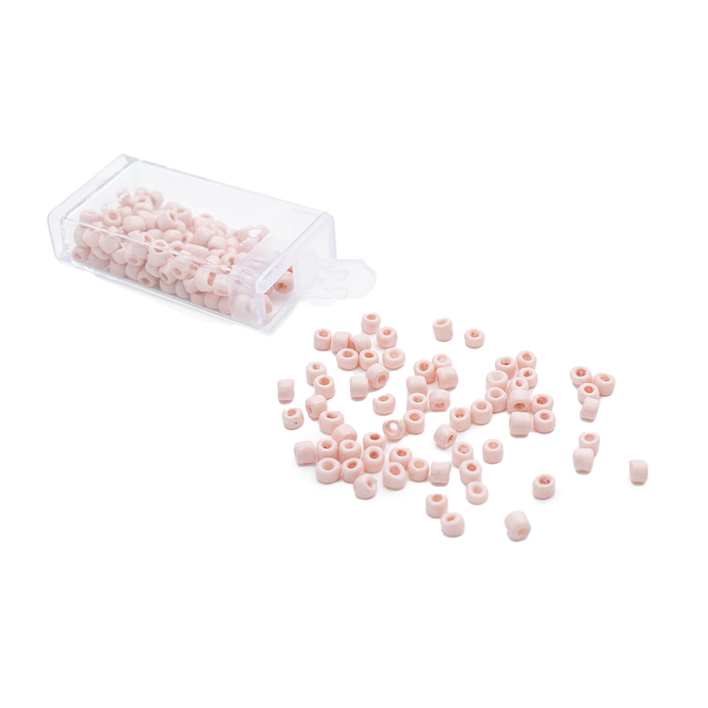 Kleine rosa Rocailles-Perlen von Alexandra Renke im Vintage-Stil liegen verstreut auf einem weißen Hintergrund neben einem durchsichtigen Plastikbehälter.