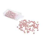 Kleine rosa Rocailles-Perlen von Alexandra Renke im Vintage-Stil liegen verstreut neben einem durchsichtigen Plastikbehälter auf weißem Hintergrund.