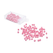 Ein durchsichtiger Plastikbehälter mit kleinen rosa Rocailles-Perlen von Alexandra Renke, von denen einige auf einem weißen Hintergrund verstreut sind – ideal, um Ihrem handgefertigten Schmuck einen Vintage-Look zu verleihen.