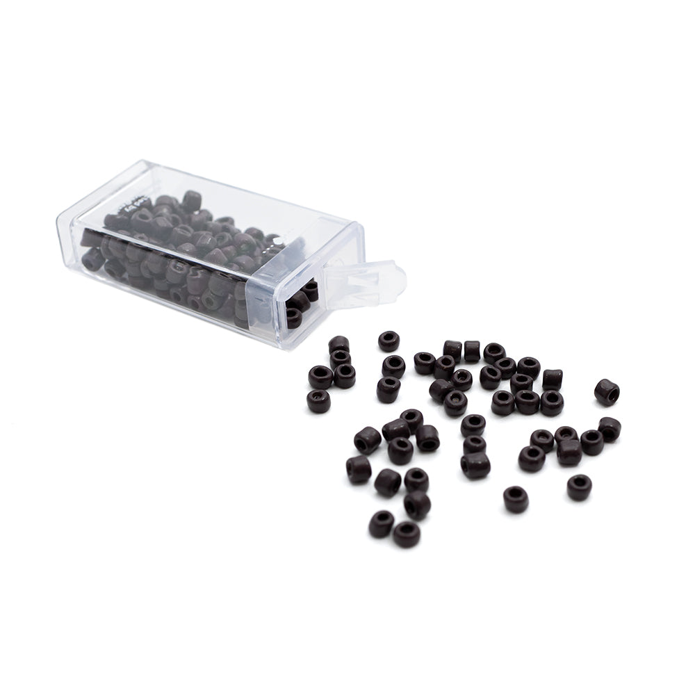 Kleine schwarze Rocailles-Perlen von Alexandra Renke rieseln aus einem durchsichtigen Plastikbehälter auf eine weiße Oberfläche – ideal, um Ihren Bastelarbeiten einen Hauch von Vintage-Stil zu verleihen.