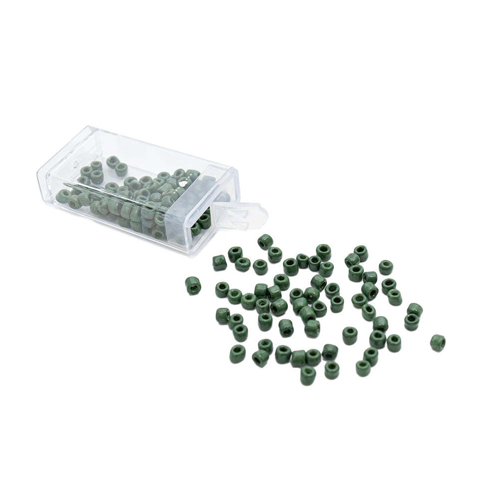 Kleine grüne Rocailles-Perlen von Alexandra Renke im Vintage-Look ergießen sich aus einem durchsichtigen Plastikbehälter auf eine weiße Fläche.