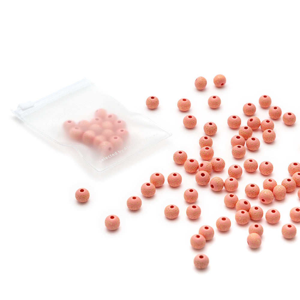Kleine rosa Holzperlen von Alexandra Renke sind auf einer weißen Fläche verstreut, einige Perlen befinden sich in einem transparenten Ziplock-Beutel.