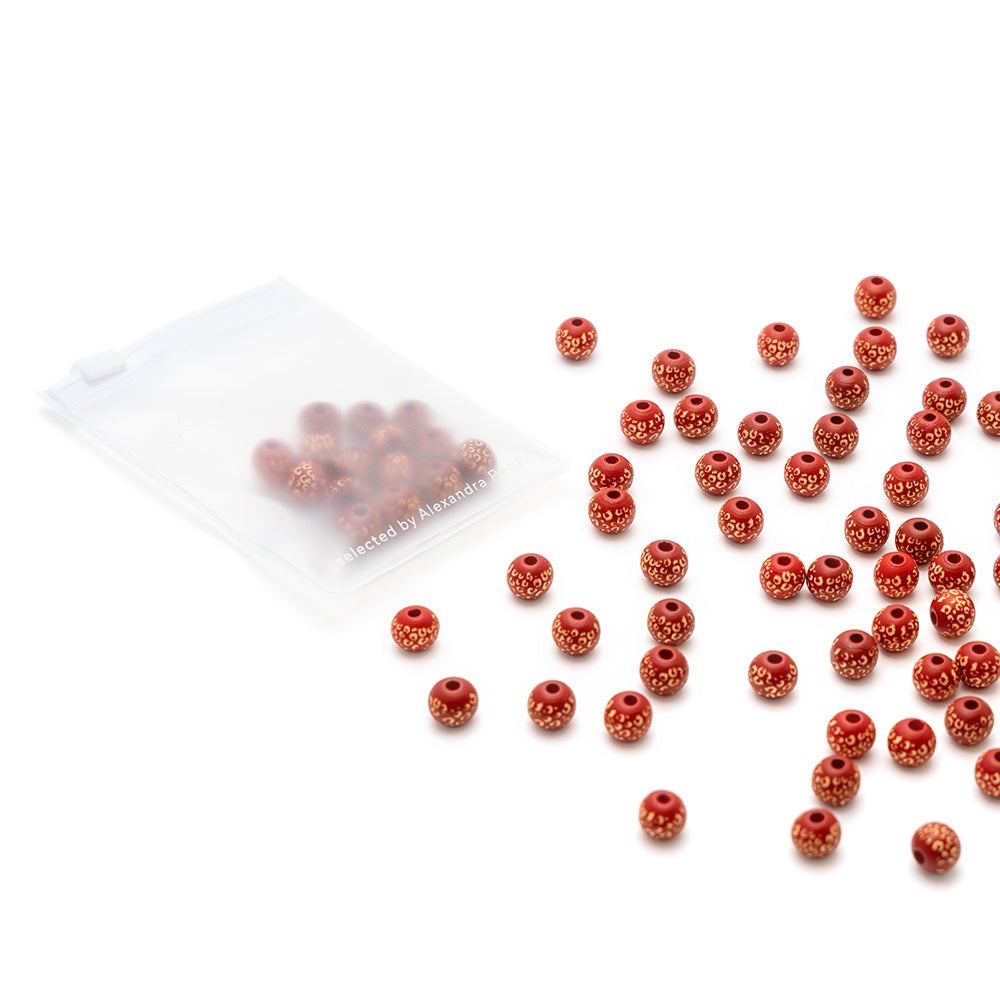Rote und goldene runde Perlen, darunter Alexandra Renke handgefertigte Holzperlen, liegen verstreut neben einem durchsichtigen Plastikbeutel, der mit weiteren Perlen gefüllt ist.